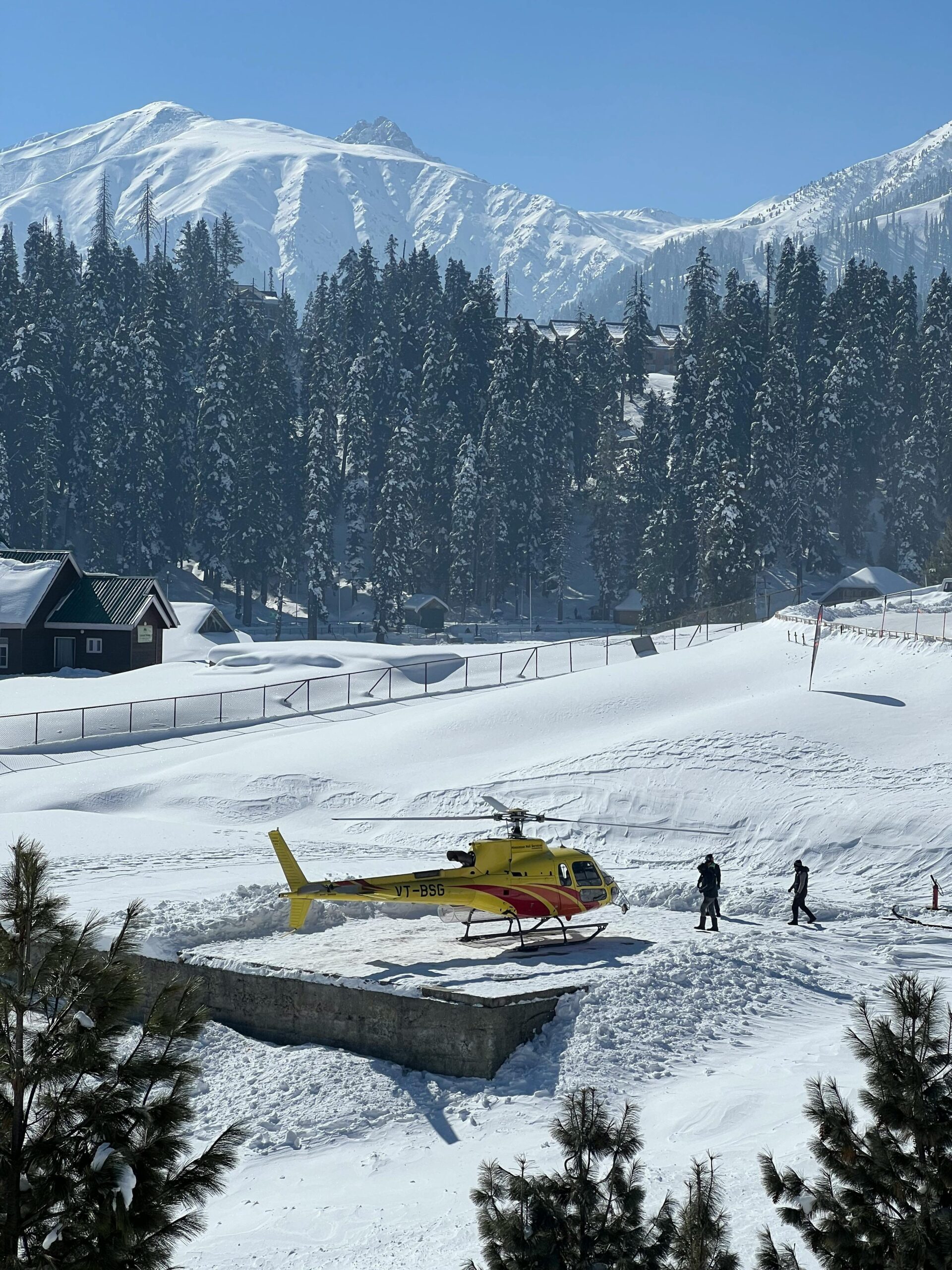 Yellow helicopter parked on snowy Gulmarg backdrop with majestic mountains and pine trees.